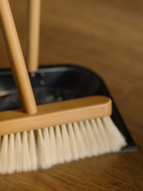 A clean, light blue dustpan and a wooden-handled scrub brush with beige bristles hang on a plain, light-colored wall. The dustpan has a smooth surface with minimal dust or debris visible, and the scru