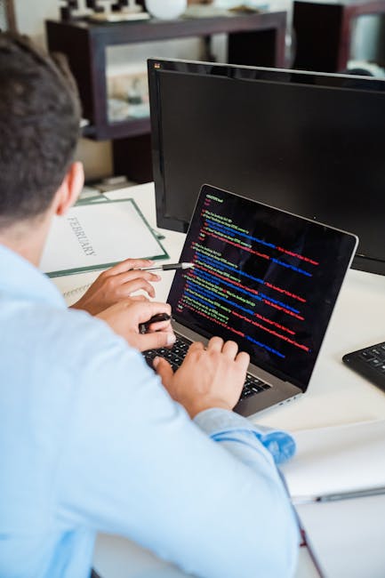 A person wearing a light blue shirt is seated at a white desk in an office environment, working on a laptop displaying lines of colorful programming code. The desk includes a black keyboard, a black m